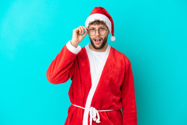 Young man disguised as Santa Claus isolated on blue background with glasses and surprised