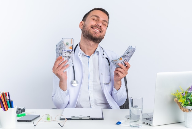 Молодой человек-врач в белом халате и со стетоскопом держит деньги счастливыми и взволнованными, сидя за столом с ноутбуком на белом фоне