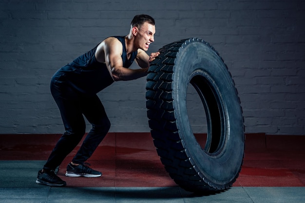 Young man doing exercises with a tire wheel in the gym on a dark background