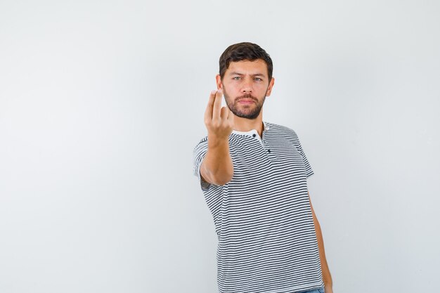 Young man doing money gesture in striped t-shirt and looking serious. front view.