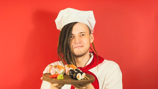 Young man dressed as chef holding wooden board with sushi rolls Male cook with appetizing sushi rolls on red background Concept of fast food and takeaway