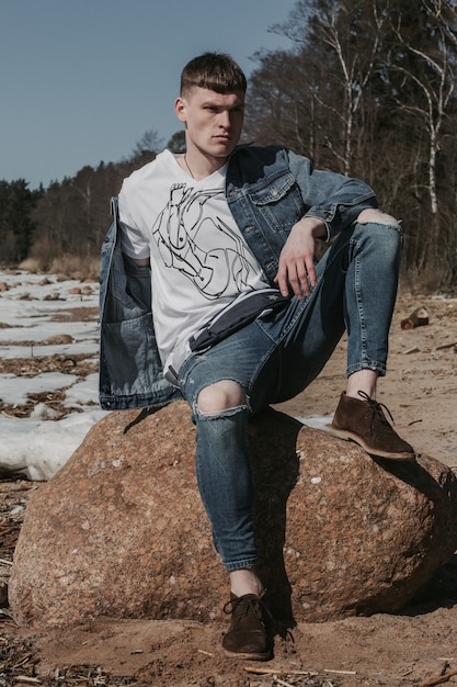 Young man dressed in casual jeans outfit posing on a stone by the cold sea shore.