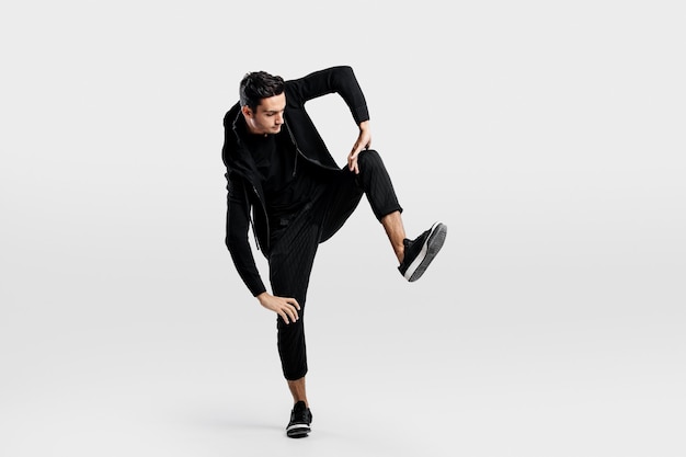 Young man dressed in a stylish black clothes lifts one leg up while dancing street dance