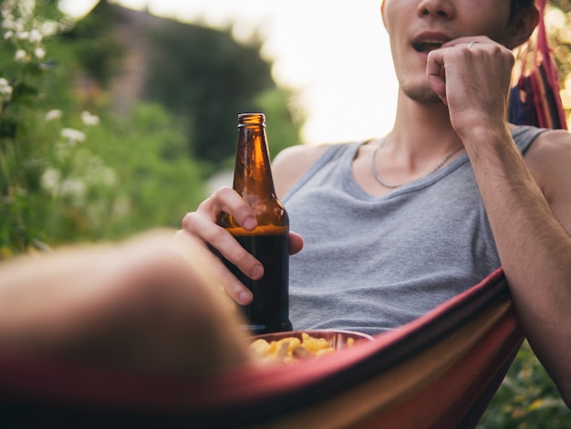 Foto un giovane che beve birra e che mangia i cracker che riposano su un'amaca in estate