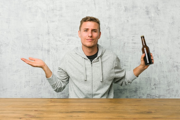 Young man drinking a beer on a table doubting and shrugging shoulders in questioning gesture.