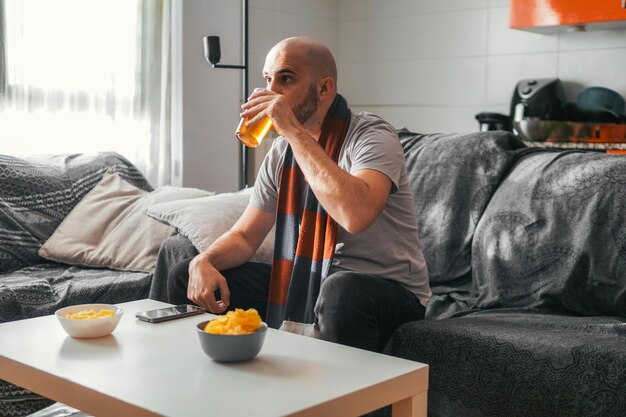 Photo young man drinking beer while watching sports at home with a blue and red sports scarf