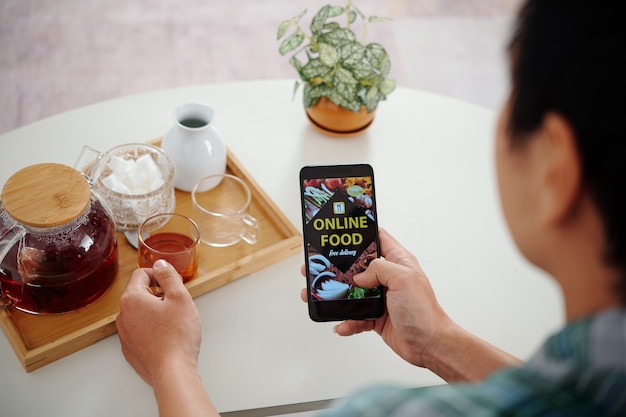 Young man drinking black tea and ordering food delivery via application on smartphone