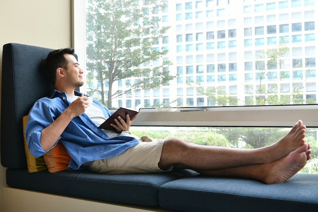 Young man drinking coffee while sitting on sofa at home with the book in his hand 