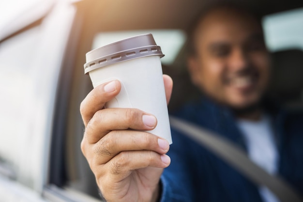 Young man drinking a cup of hot coffee while driving car to\
travel hands holding steering wheel take a break for a refreshing\
coffee before your trip