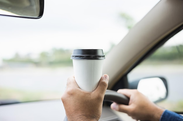 Young man drinking a cup of hot coffee while driving car to\
travel hands holding steering wheel