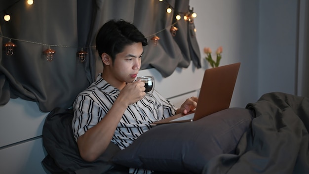 Young man drinking hot tea and laptop computer on bed at night.