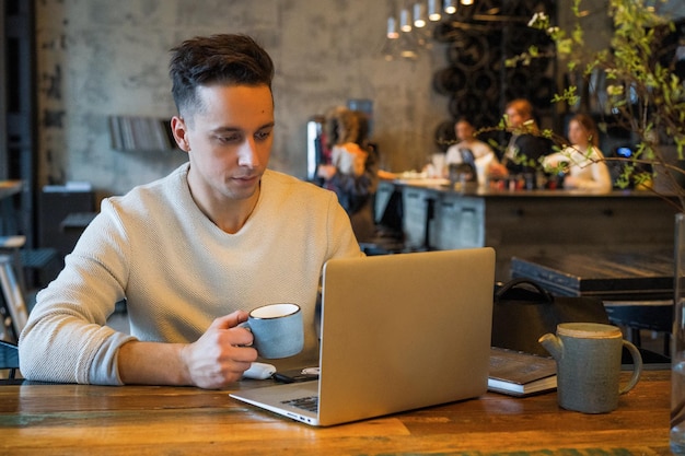 young man drinks tea and works on a laptop, freelancer working in a cafe