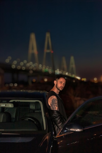 Young man driver standing by car at night thoughtfully looking at camera Sad depressed guy alone thinking about problems feeling lonely