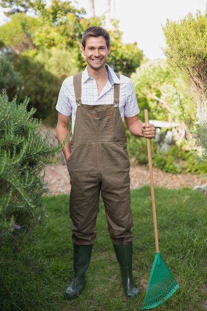 Молодой человек в dungarees проведение грабли в саду