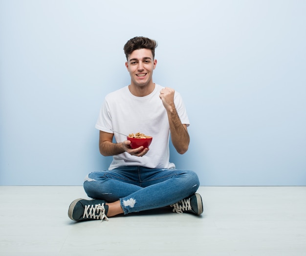 Young man eating cereals sitting on the floor cheering carefree and excited. Victory concept.