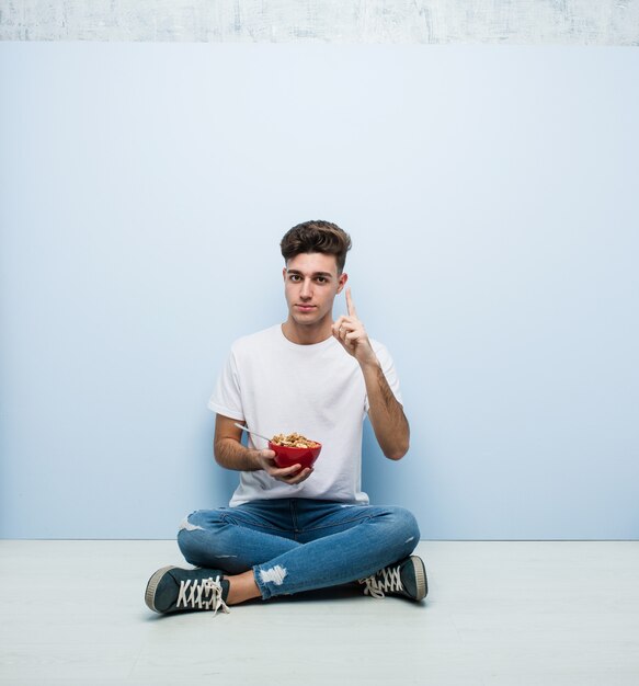 Young man eating cereals sitting on the floor showing number one with finger.