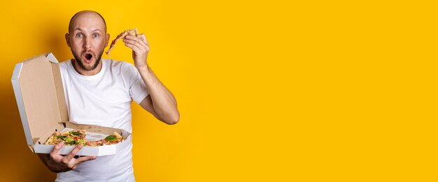 Young man eating hot fresh pizza with packaging on a yellow surface. Banner.