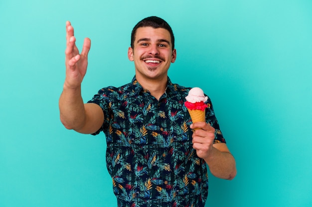 Young man eating an ice cream isolated on blue wall receiving a pleasant surprise, excited and raising hands