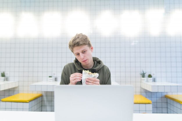 Un giovane mangia un panino e usa un laptop in un caffè leggero.