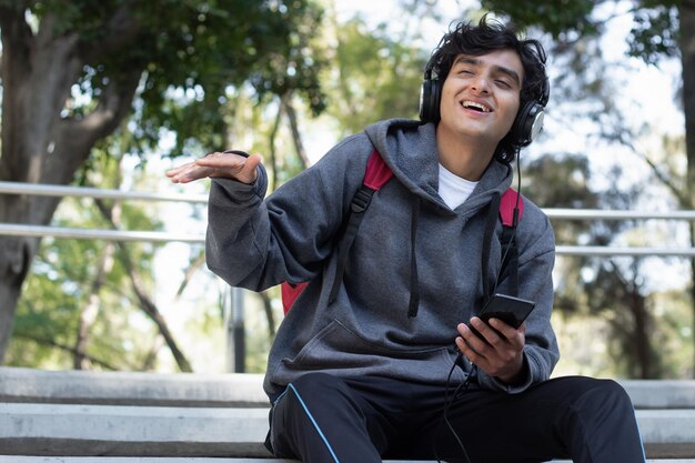 Young man enjoying some time alone relaxing listening to music with his headphones in the park, he has a smile on his face and a facial expression of peace and well-being.