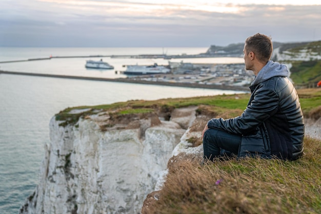 Young man exploring the White Cliffs of Dover in UK. Sitting on top of the cliff watching English canal.