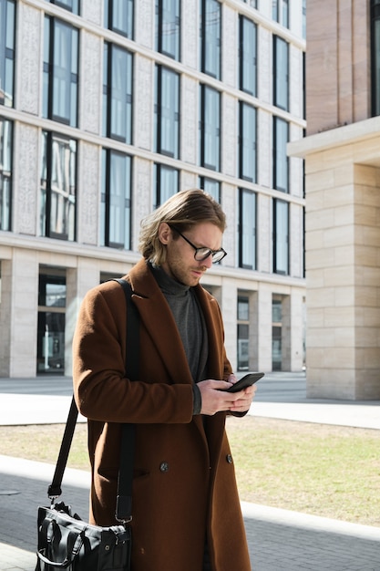 Young man in eyeglasses using mobile phone to find a way while walking along the street in the city