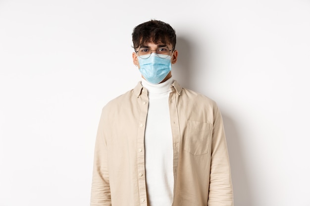 Young man in face mask from covid-19 looking cheerful , standing on white wall. Coronavirus, quarantine and social distancing concept.