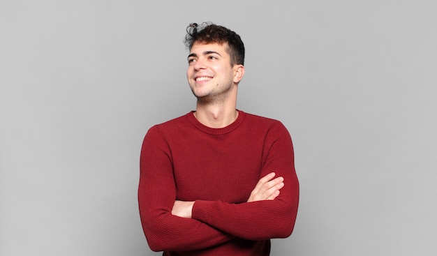 Young man feeling happy, proud and hopeful, wondering or thinking, looking up to copy space with crossed arms