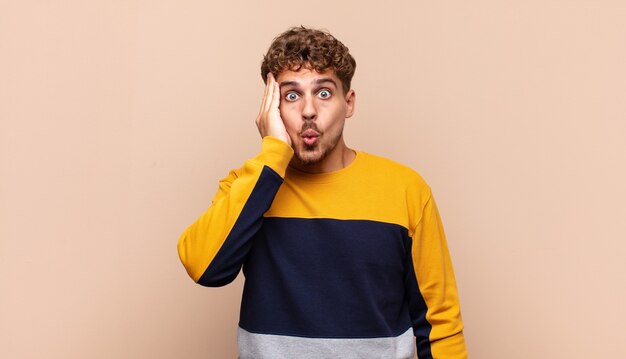 Young man feeling shocked and astonished holding face to hand in disbelief with mouth wide open