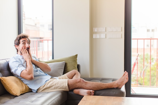 Young man feeling shocked and scared, looking terrified with open mouth and hands on cheeks