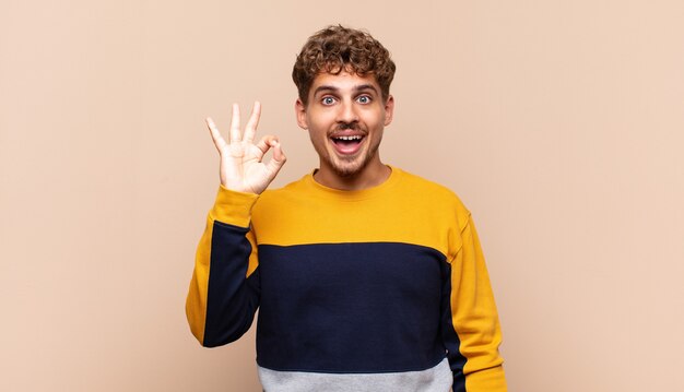 Young man feeling successful and satisfied, smiling with mouth wide open, making okay sign with hand