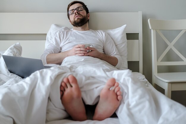 Young man fell asleep with a circle in hand sitting in bed he worked remotely all night with laptop