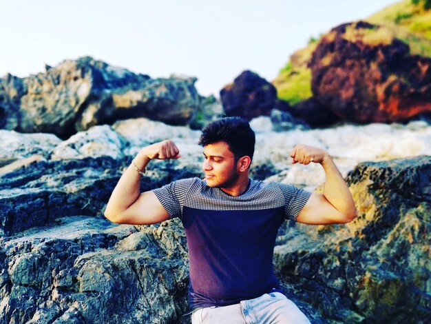 Photo young man flexing muscles while sitting on rock