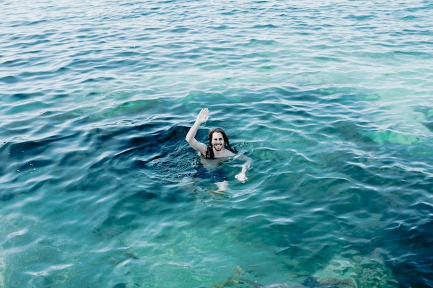 Young man floating in the ocean during his holidays in spain, crystal clear sea, saluting to the camera, copy space