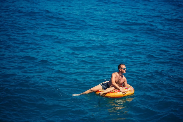 한 젊은이가 푸른 물이 있는 바다의 팽창식 에어 링 원 위에 떠 있습니다. 행복한 화창한 날에 축제 휴가