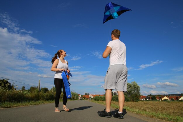 Young man flying kite by smiling girlfriend on road during sunny day