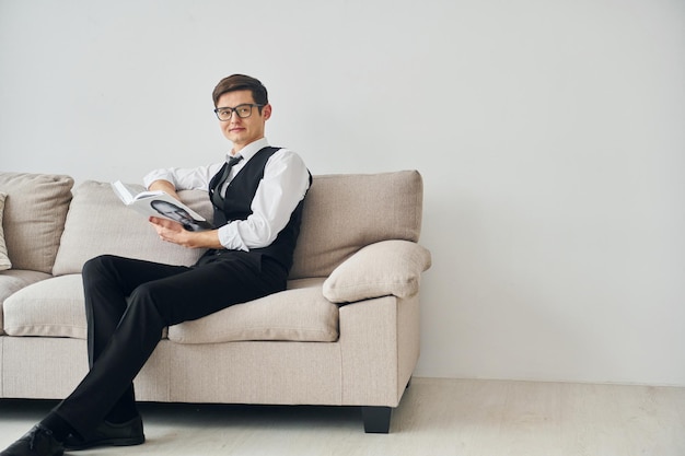 Young man in formal clothes sits on the sofa indoors against white wall