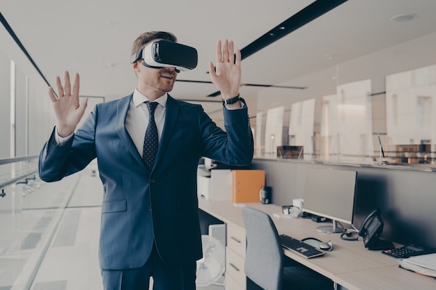 Young man in formal suit enjoying virtual tour or excursion with VR headset. Businessman in virtual reality glasses standing near working desk in office, tries to touch objects in digital actuality