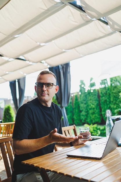 Young man freelancer developer enjoying coffee break on summer terrace of cozy cafe working remotely on laptop smiling looking at laptop