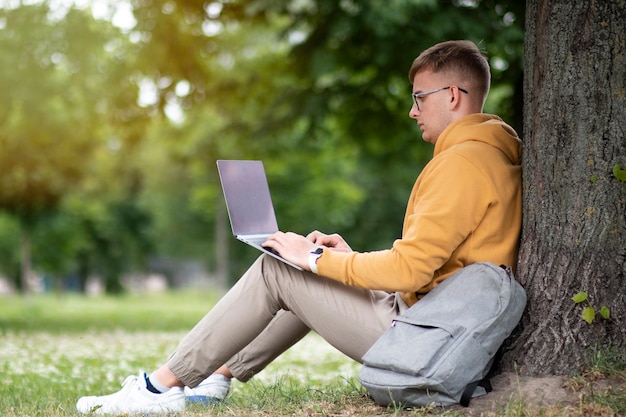 Young man freelancer is working on laptop in glasses leaning on a tree in a park