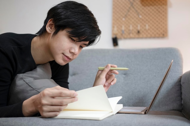 Young man freelancer making note on notebook and using laptop on sofa