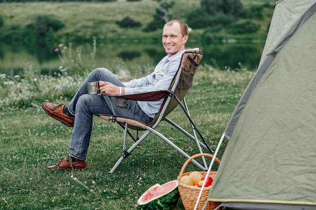 Young man freelancer sitting on chair and relaxing in front of tent at camping site in forest or meadow. Outdoor activity in summer.