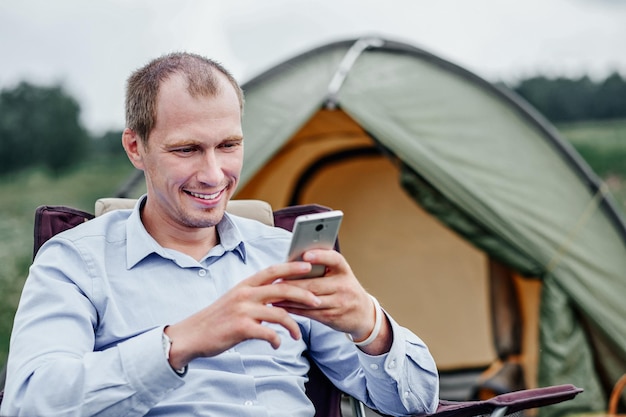 Giovane libero professionista seduto su una sedia e utilizzando lo smartphone rilassarsi davanti alla tenda in campeggio nella foresta o nel prato lavoro a distanza e attività all'aperto in estate