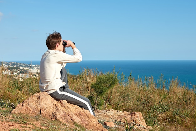 A young man from Spain in a white shirt sitting on a rock looking over the sea through binoculars