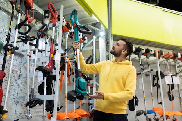 A young man in a gardening equipment store chooses an electric trimmer for mowing lawn grass