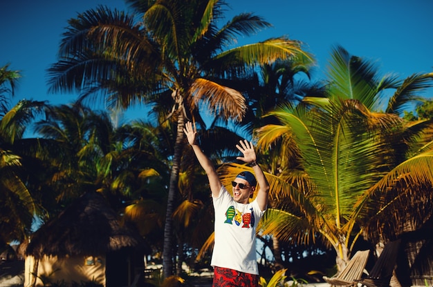 Photo young man gesturing triumph with raised hands against palm trees background