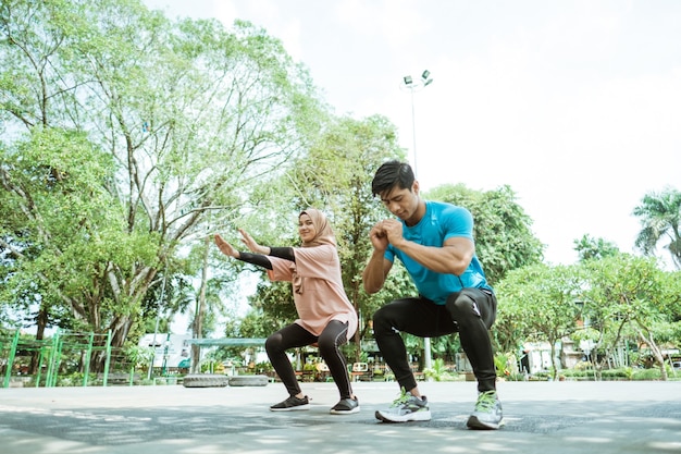 A young man and a girl in a headscarf doing squat movements together when outdoor exercise in the park