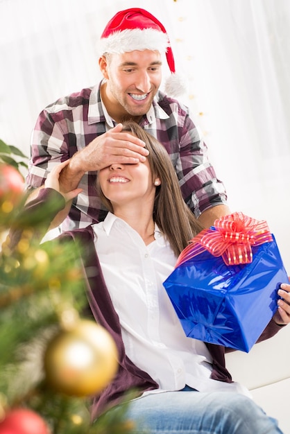 젊은 남자는 그의 여자 친구에게 그녀의 눈 위에 손을 잡고 크리스마스 선물을 줍니다.