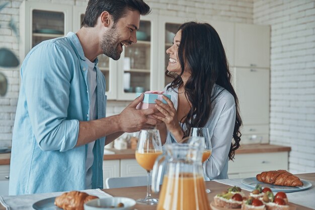 Young man giving a gift box to his girlfriend while having breakfast at the domestic kitchen