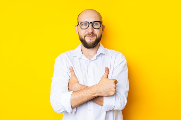 Young man in glasses arms crossed holding thumbs up on yellow background